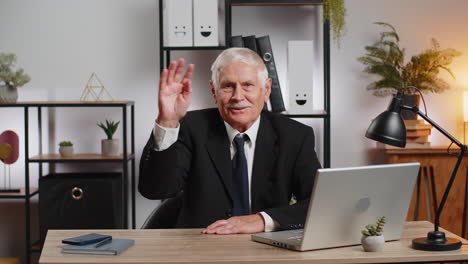 happy senior businessman with laptop waves hand hi gesture greeting welcomes someone at home office