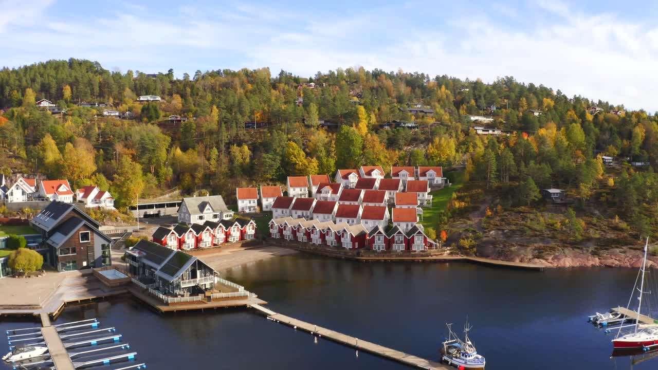 The drone soars over Holtnesstøa's port village, closing in on vibrant houses by calm waters. Autumn hues adorn the scene, as the colorful rooftops stand out against the seasonal backdrop.