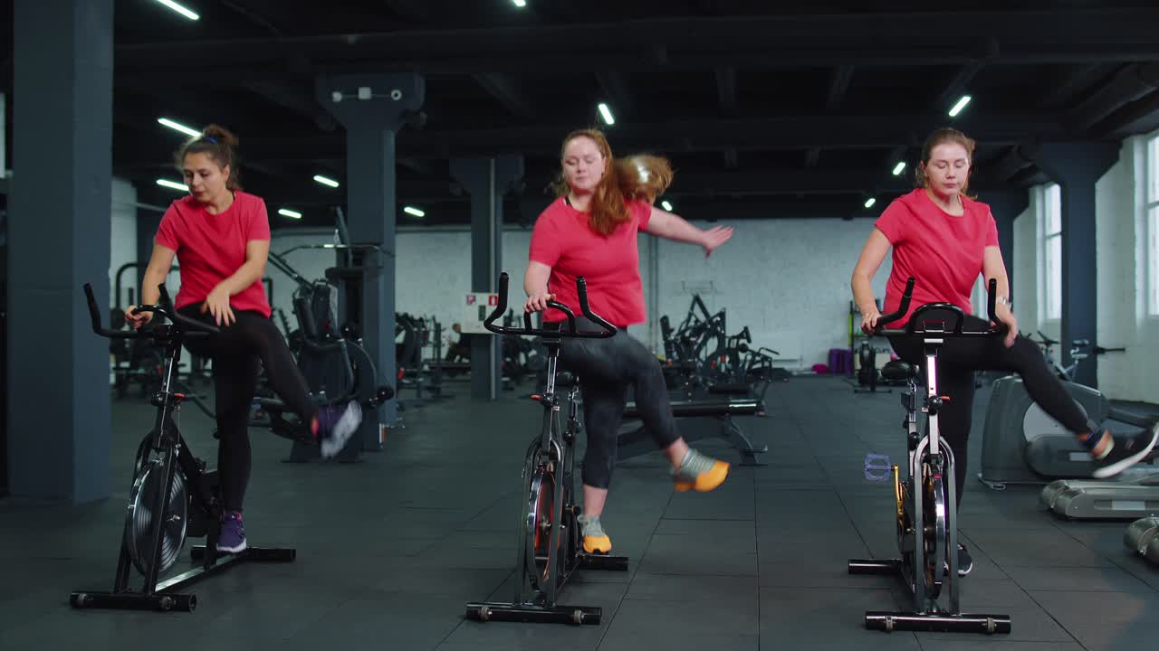 건강한 백인 여성 그룹이 체육관에서 정지 자전거 기계 자전거에서 운동을합니다.