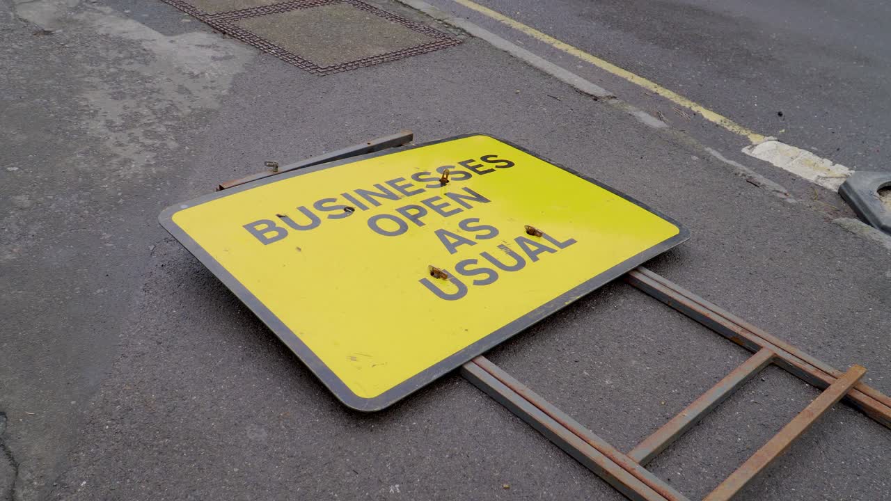 Close-up shot in Cheddar of a bright yellow roadside sign reading “Business Open As Usual,” laying beside a quiet street, with village buildings softly blurred in the background as the camera gently