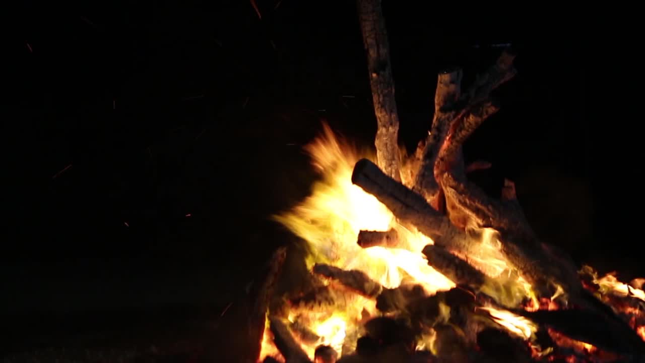 4K close-up of glowing bonfire at night. Bright flames, wood crackling and flying sparks create cozy and intense camping vibe. Perfect for cinematic, nature or travel themes.