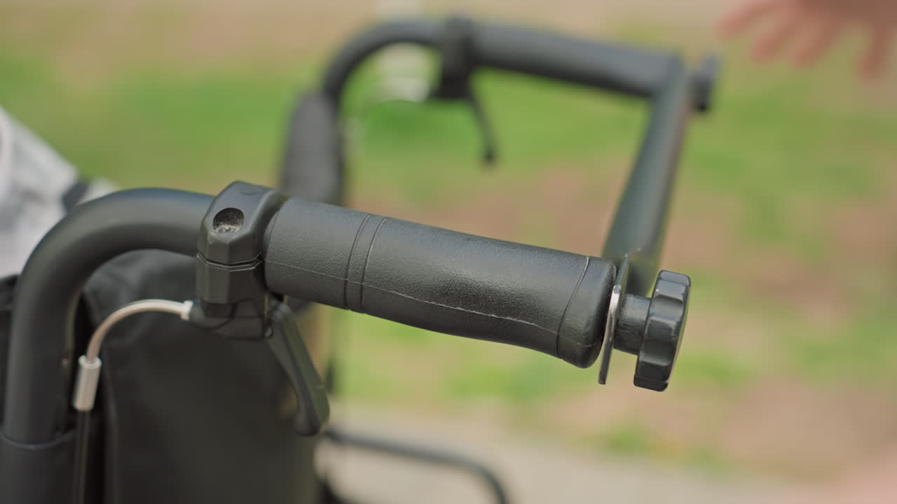 Hand Adjusting Wheelchair Brake Lever Closeup On Handle And Cable, Tactile Focus On Lever Mechanism And Grip, Outdoor Park Sidewalk Background, Safety And Maintenance Tone In Candid Care Scene