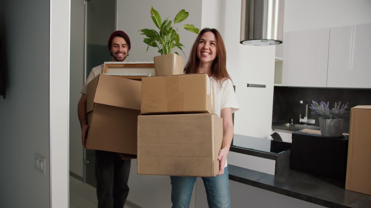 una pareja feliz una chica morena con una camiseta blanca y su novio moreno con paja en una camisetta crema traen cajas de cosas a su nuevo apartamento moderno y ponen las cajas en el sofá que está cubierto con una cubierta de plástico