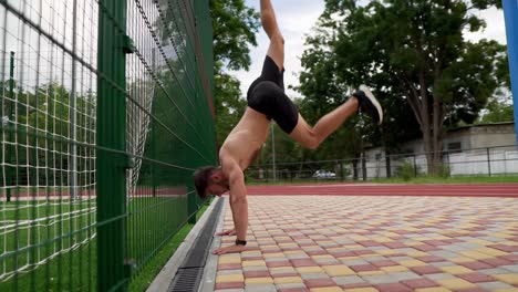 The-young-athlete-is-doing-pushups-on-hands-standing-upside-down-near-the-wall-outdoors.-Young-man-topless-in-black-shorts