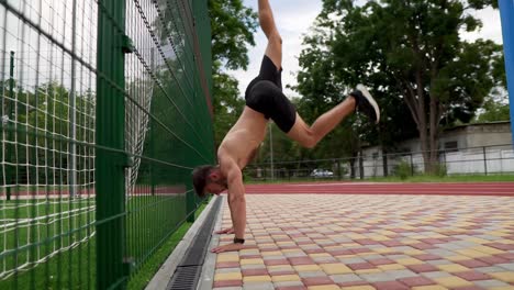 the young athlete is doing pushups on hands standing upside down near the wall outdoors. young man topless in black shorts