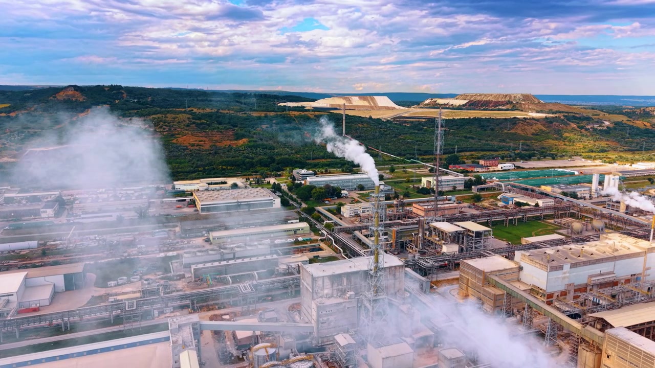 Industrial chemical plant structures with vapor rising from exhaust pipes. The aerial view captures complex pipeline systems and processing units next to green hills under cloudy skies.