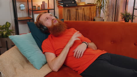 happy young man in wireless headphones relaxing lying on sofa at home listening favorite music