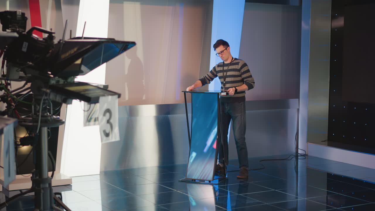 man adjusts digital display on podium in video production studio, working with equipment, camera setup, lighting, blue background, preparing studio set for filming, focused on screen configuration
