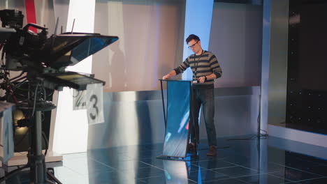 man adjusts digital display on podium in video production studio, working with equipment, camera setup, lighting, blue background, preparing studio set for filming, focused on screen configuration