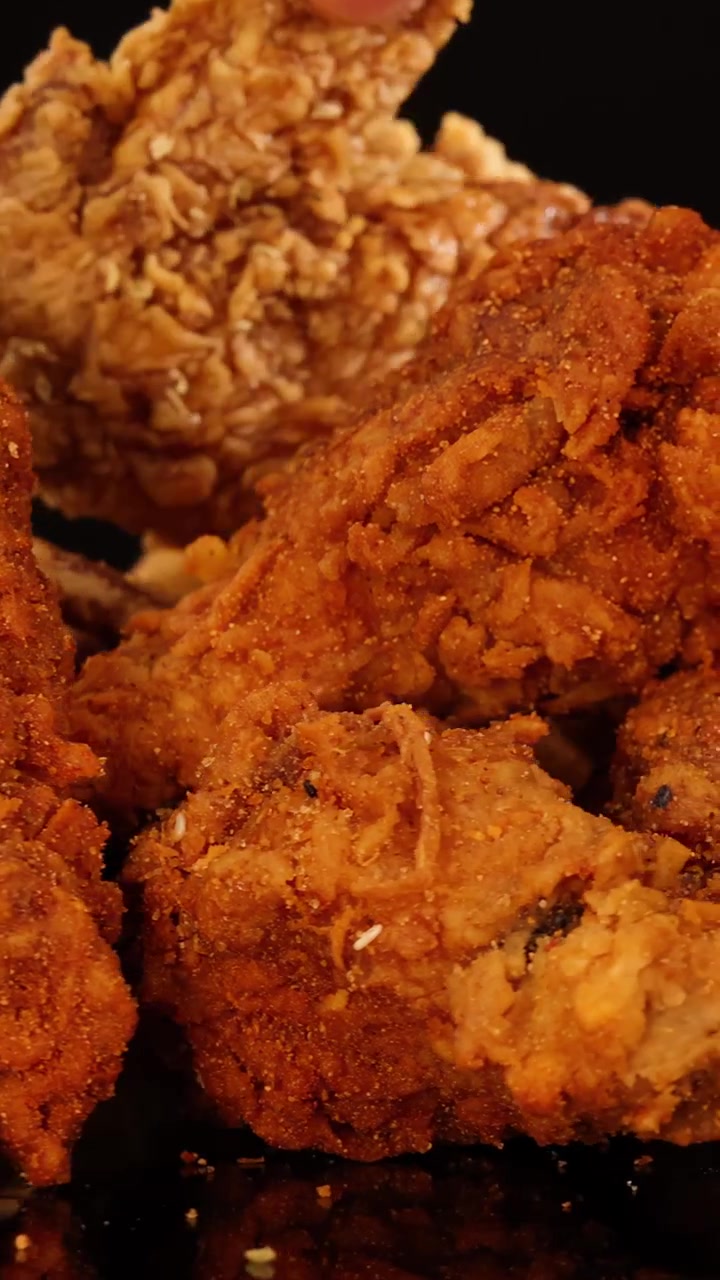 A hand reaches in to pick up assorted crispy fried chicken wings, drumsticks, and nuggets under warm studio lighting, with a dark, minimal backdrop