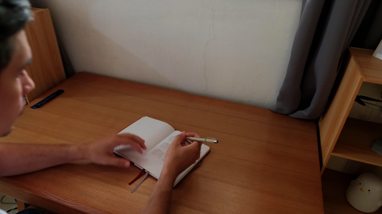 Static overhead medium shot of young male student writing in journal notebook atop wooden desk