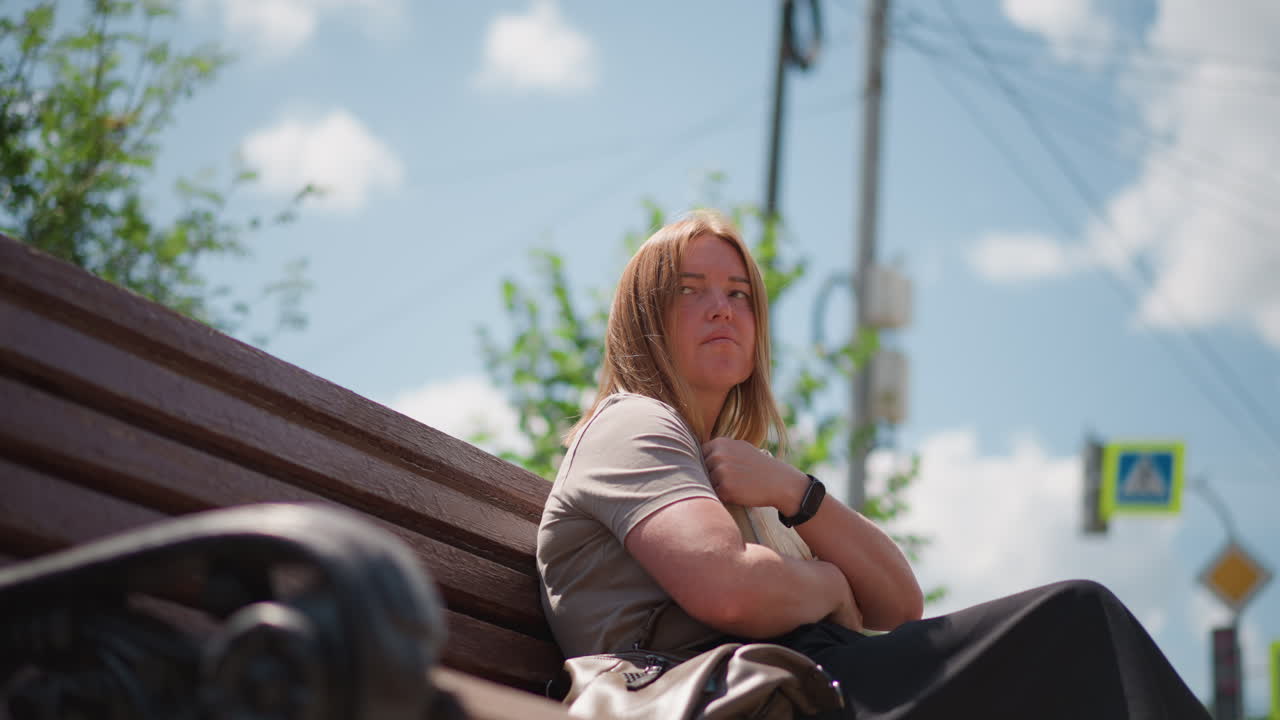 Calm lady sits on wooden bench under warm sunlight flipping book pages then gently closes it and presses it to chest while pigeon flies nearby surrounded by green trees