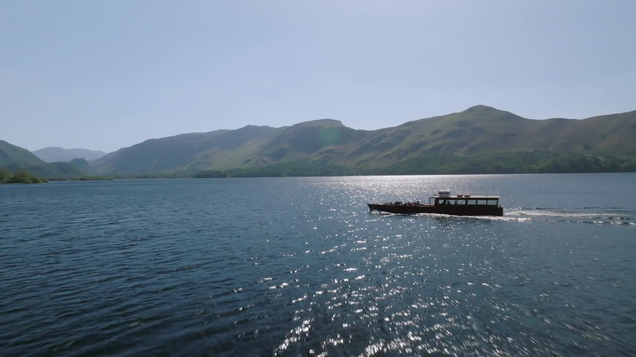 Flying towards and over small ferry boat moving on Cumbrian lake with distant fells. Derwentwater, Lake District, Cumbria, UK.