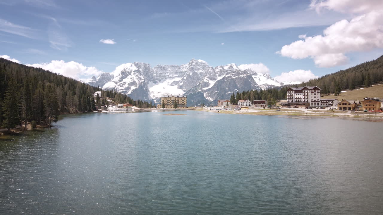 Camera flies straight ahead over a sparkling alpine lake toward a grand lakeside hotel and clustered lodges, framed by dense forest and towering snow-covered peaks