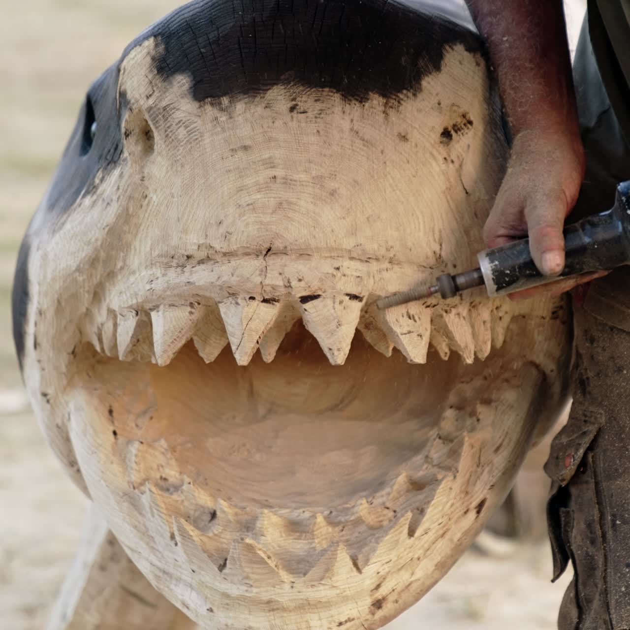 Unrecognized craftsman making a wooden sculpture of a shark. Artist uses a grinding tool to cut sharp teeth of a fish