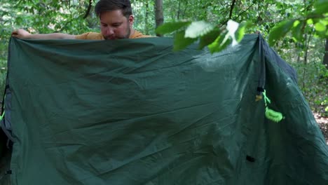 young adult man packing up tent after sleeping in forest, handheld view