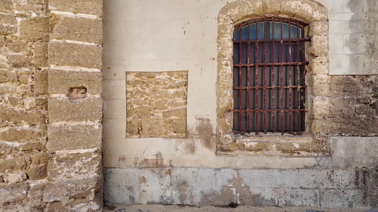 Vista ravvicinata di un antico muro in pietra con una porta di legno e una finestra con inferriate su un edificio storico a Cadice, Spagna. Le texture consumate e l'architettura tradizionale riflettono il ricco patrimonio della città