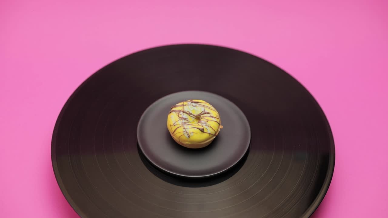 An American donut topped with yellow chocolate with blue and black choco lines turns in a circle on a black turntable. The shot from above