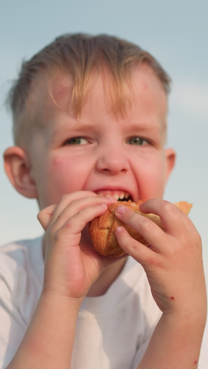 A little boy in a white shirt sits in a grassy field, carefully holding a snack with both hands. He gazes at it intently before taking a bite, enjoying a peaceful moment outdoors in the warm sunshine