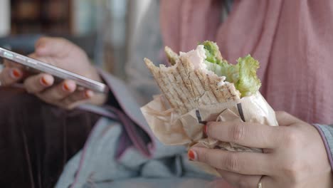 woman eating sandwich while using phone in cafe