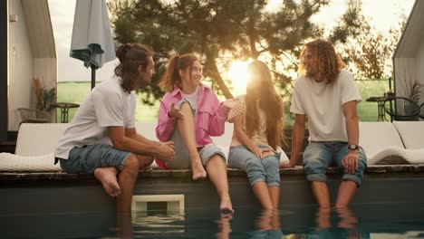 four friends sit near the sunbeds by the pool and have fun chatting with each other against the background of the sunset. rest in the country house