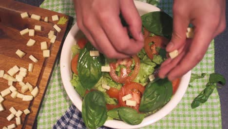 hands making a lettuce and tomato salad with basil and olive oil, adding a touch of cheese, to the mixture in the white bowl