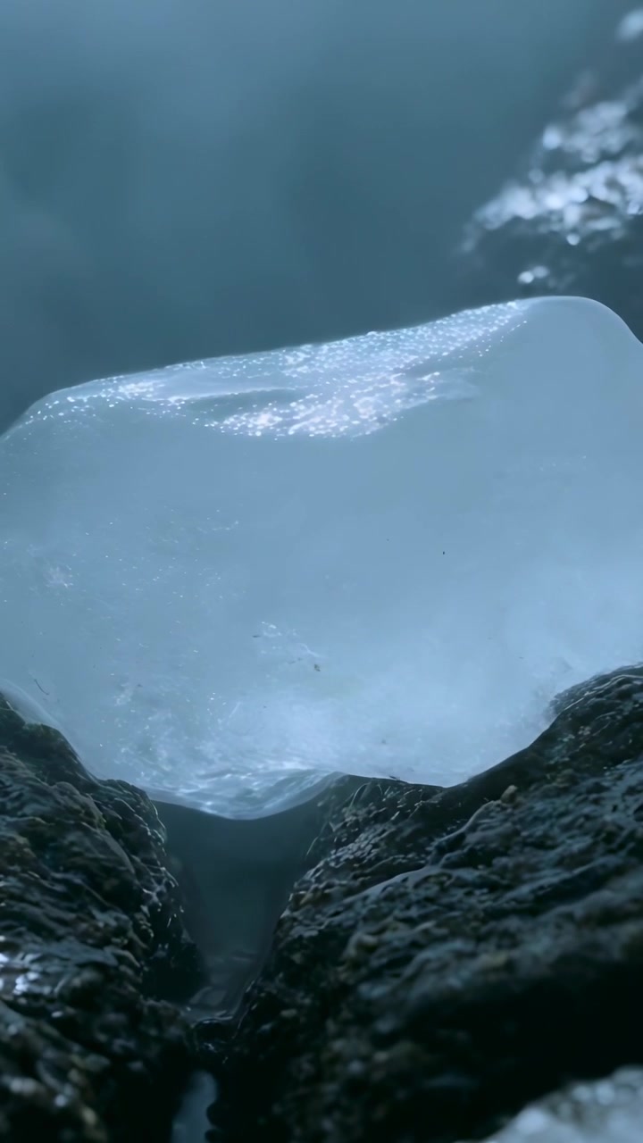 Video verticale: Telecamera che si avvicina, un pezzo di ghiaccio blu che si scioglie tra rocce scure e bagnate, formando un ruscello.