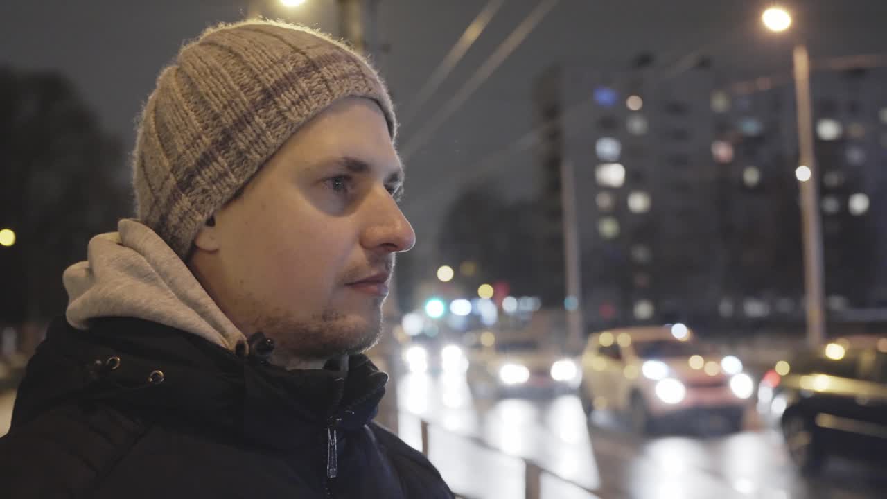 задумчивый молодой человек стоит на улице в городе в холодную ночь с движением транспорта, с ручной тележкой наперевес