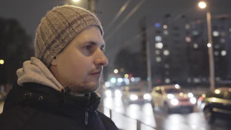 Thoughtful-Young-Man-Standing-on-a-Street-in-City-on-a-cold-Night-with-traffic,-handheld-dolly-out