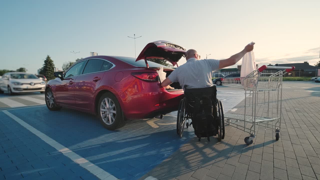 체어에 앉은 장애인 성인 남자가 슈퍼마켓 주차장에서 차의 트크에 물건을 넣고 있다.