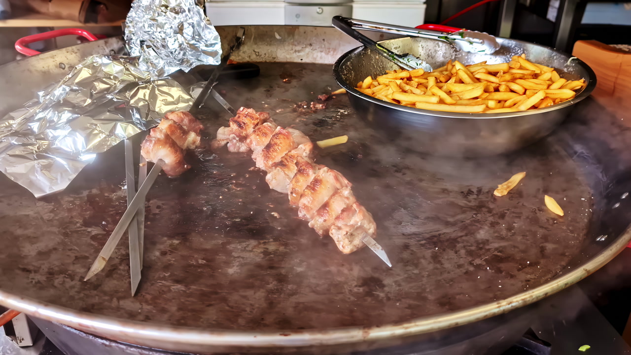 Street food cooking on a hot griddle with smoky steam, metal skewers of grilled meat and a bowl of crispy fries. Fast food prep at an outdoor stall.