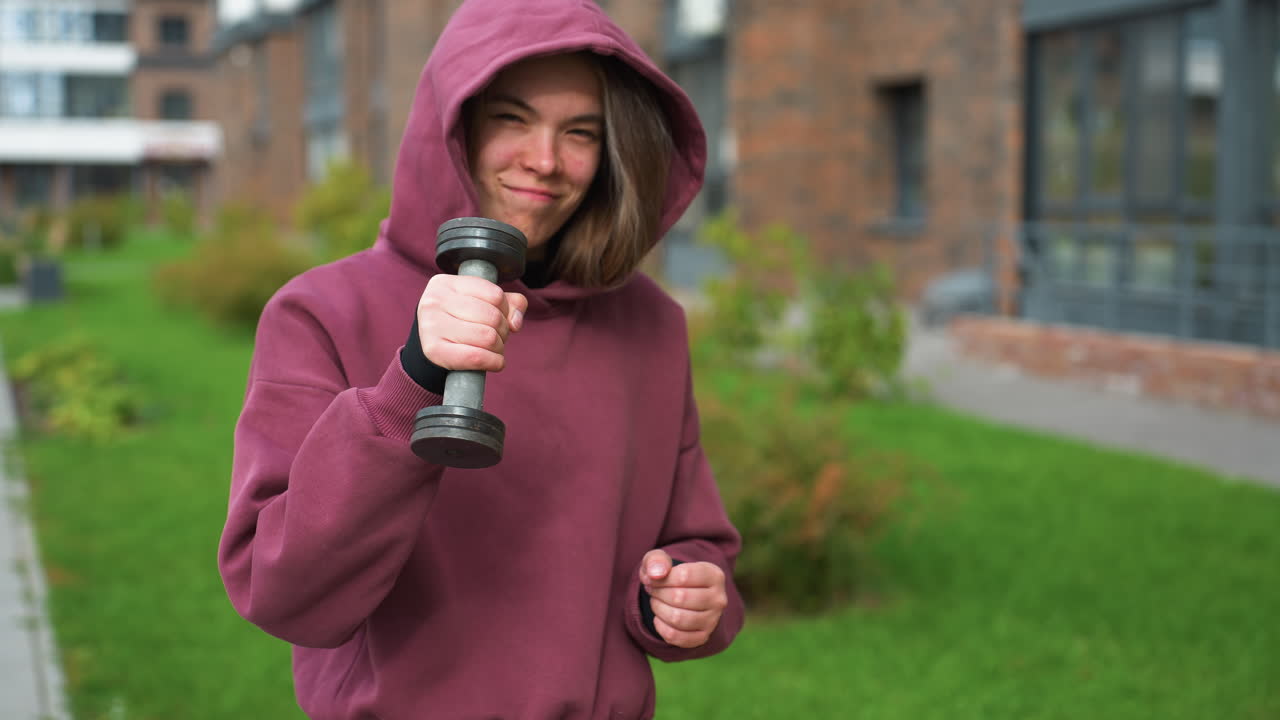 Smiling exercise enthusiast in wine hoodie confidently lifts dumbbell outdoors, showcasing strength and joy in fitness lifestyle, framed by green lawn and brick building in urban background