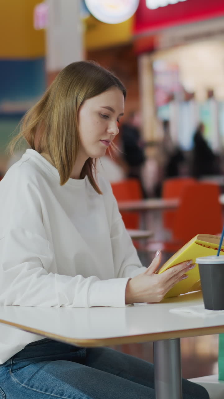 Young woman sitting in a busy mall casually reading book, surrounded by vibrant mall atmosphere, colorful shopping bags on table, and blurred background with people and fast-food counters visible