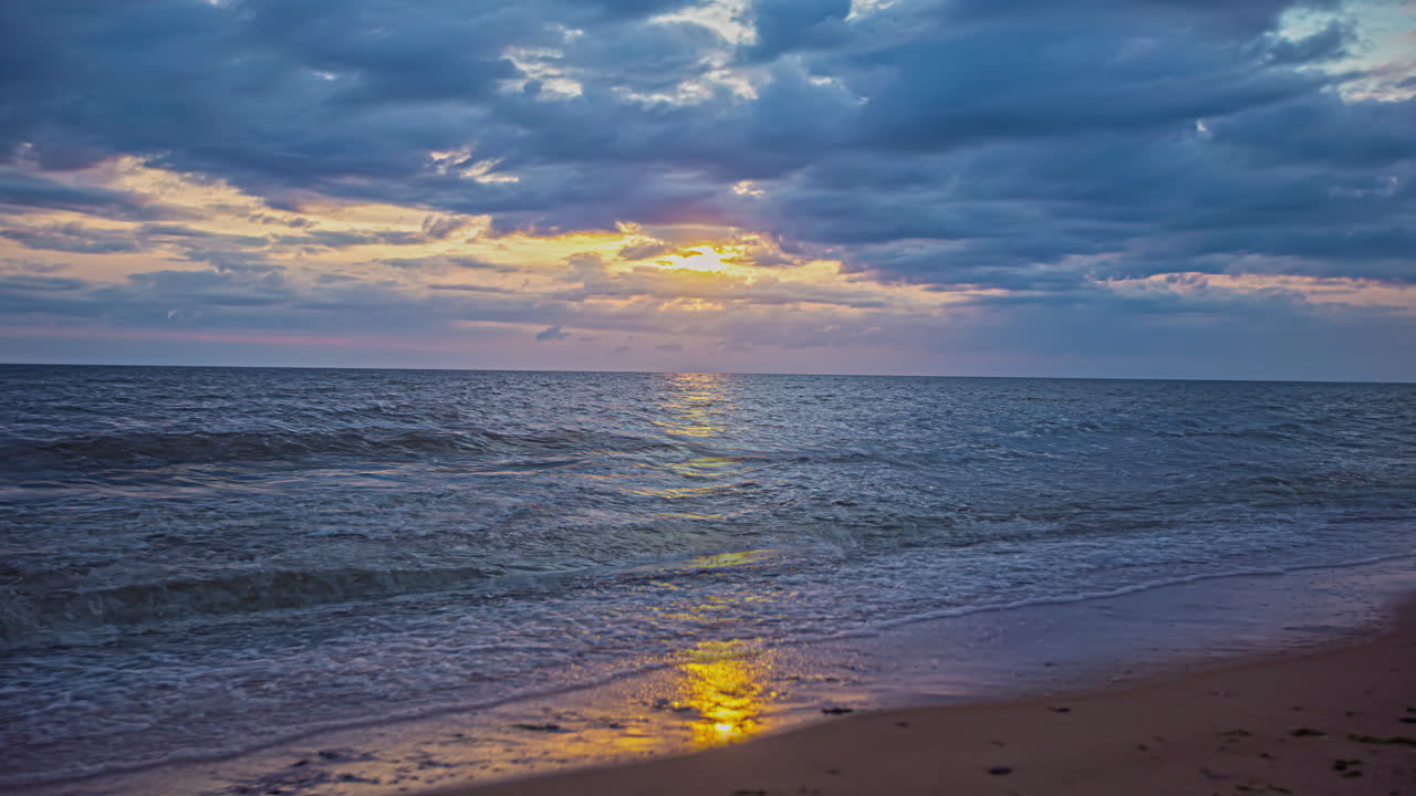 toma estática de olas tropicales que se estrellan en la playa de arena durante la noche nublada con la puesta de sol en un lapso de tiempo en el fondo