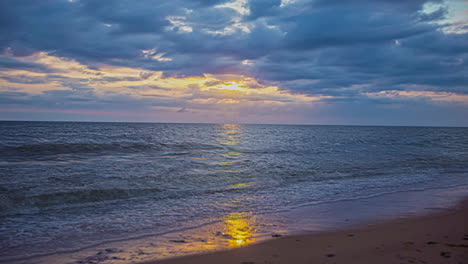 static shot of tropical waves crashing onto sandy beach during cloudy evening with sun setting in timelapse in the background
