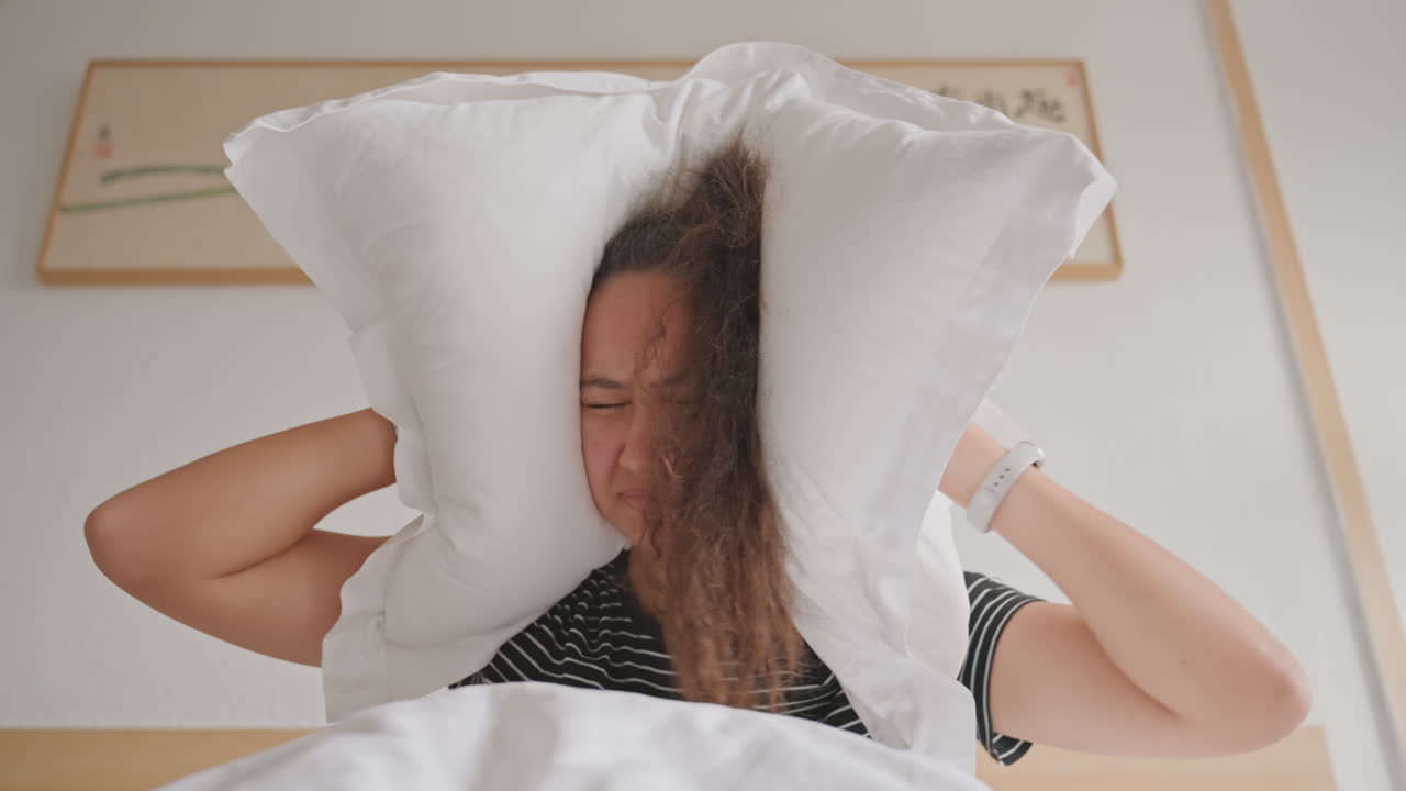 Donna rassegnata nel letto d'hotel si copre il viso. Affonda il viso sotto il cuscino e il piumino in una stanca rassegnazione, occhi chiusi, postura sconfitta, lenzuola bianche, opera d'arte sul comodino, arredamento morbido e neutro, ritratto spontaneo