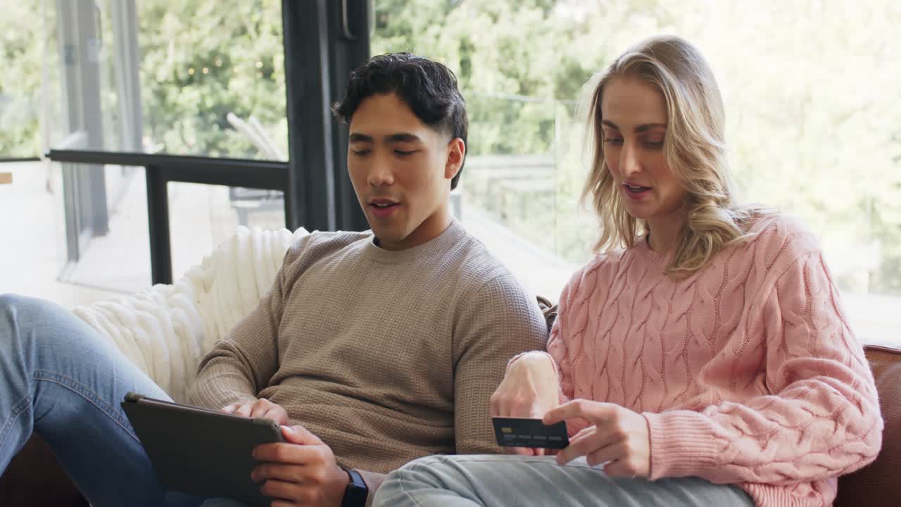 多様なカップルがソファに寄りかかり、タブレットをタップしてクレジットカード情報を取得し、取引を完了する様子