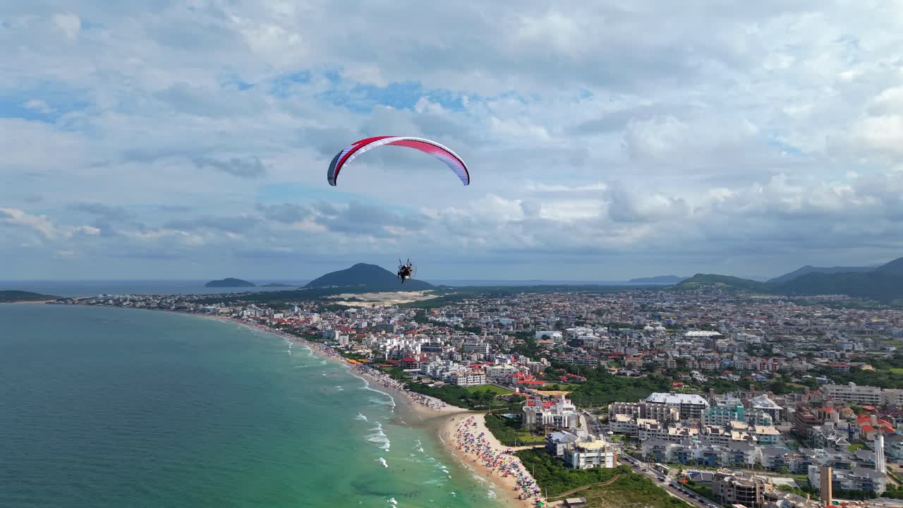 Florianopolis, Santa Catarina'daki Praia dos Ingleses üzerinde, şehir binaları, okyanus kıyısı, kum tepeleri ve yeşil dağları gösteren motorlu yamaç paraşütü uçuşunu takip eden drone çekimi.