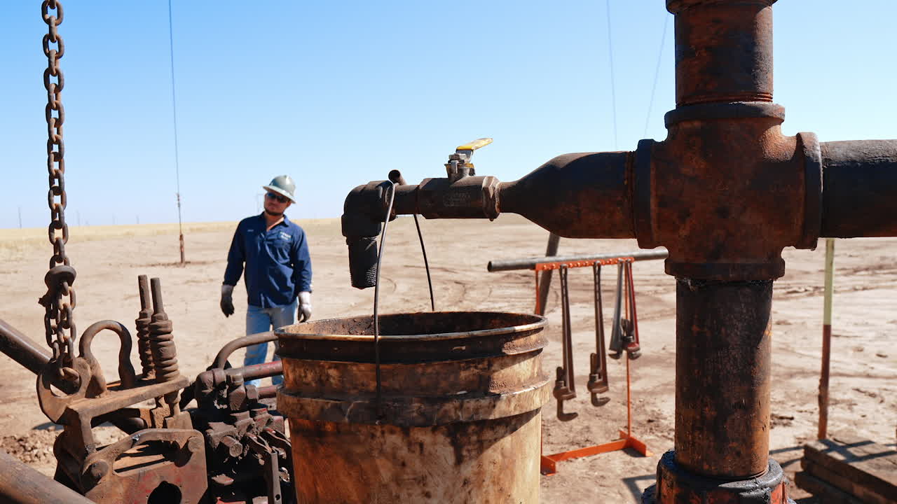 Hand of unrecognized man in a glove opening the faucet on the rusty old metal pipe. Dirty water dripping into the bucket. Worker in uniform and helmet stands at backdrop. Oil industry.