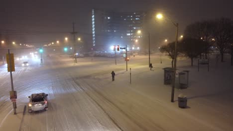 cars driving slow, on a city street covered in snow, during a snow storm at night