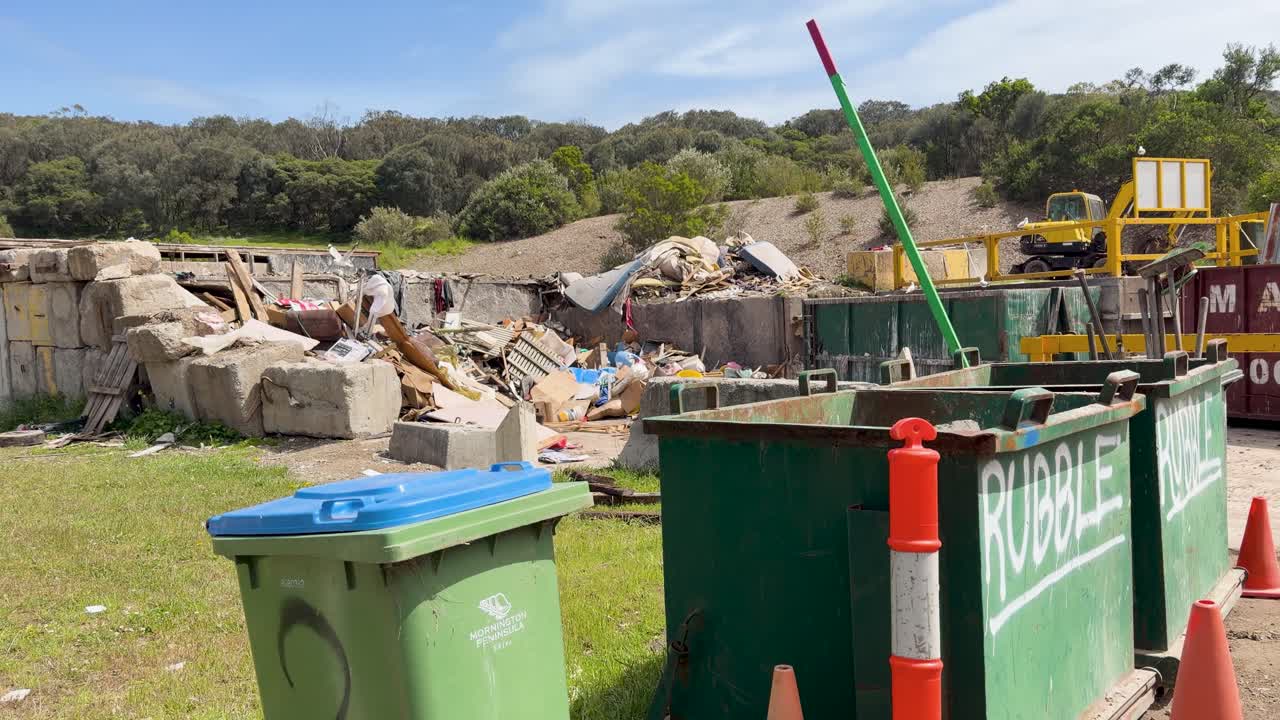 Camera slowly pans across landfill site with rubble, trash bins, and green landscape under daylight