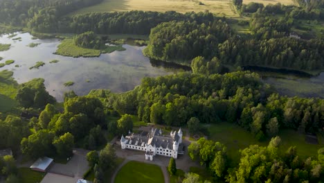 Hight altitude aerial drone footage of a white colored majestic and fairy tale like castle near a large lake and that is surrounded by a forest during sunset hour in Alatskivi. Water is reflective