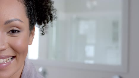 Half-portrait-of-happy-biracial-woman-in-bathrobe-in-bathroom-with-copy-space,-slow-motion