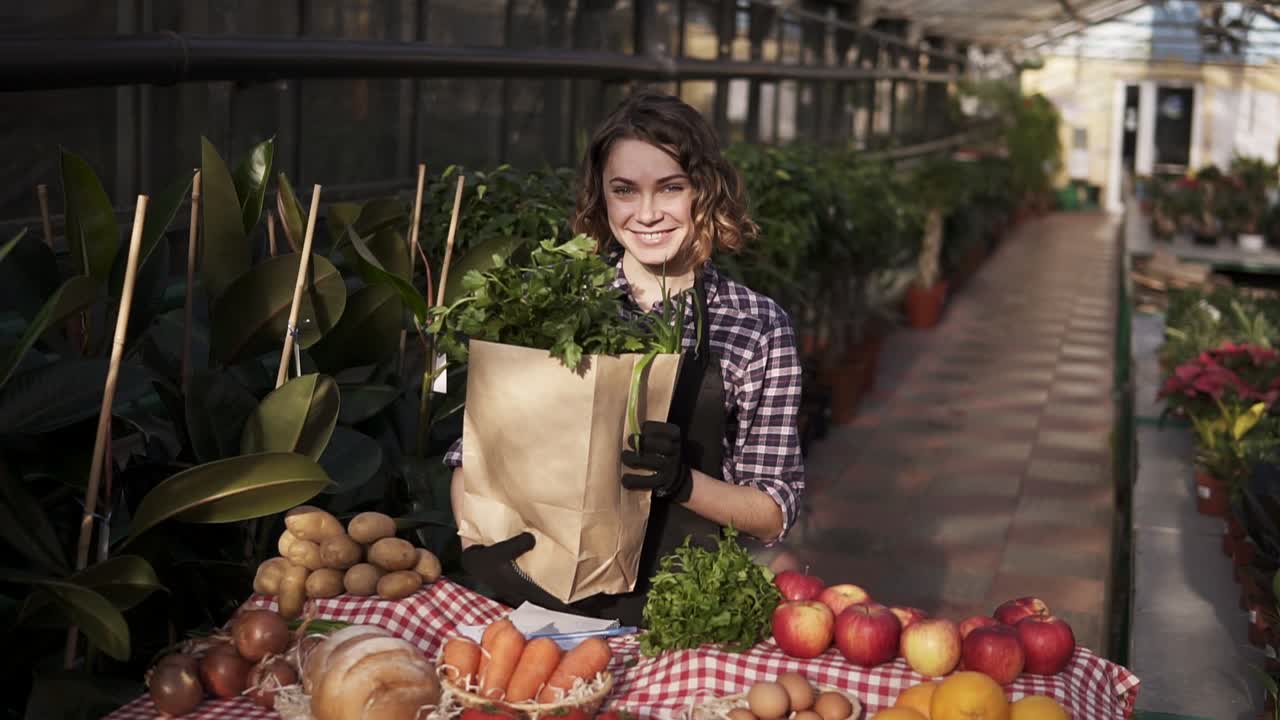 mujer joven en delantal sosteniendo una bolsa de papel marrón con verduras frescas y verduras, sonriendo mientras posa para la cámara en el mercado de alimentos de pie en una fila de invernadero interior frente a la mesa con comida orgánica