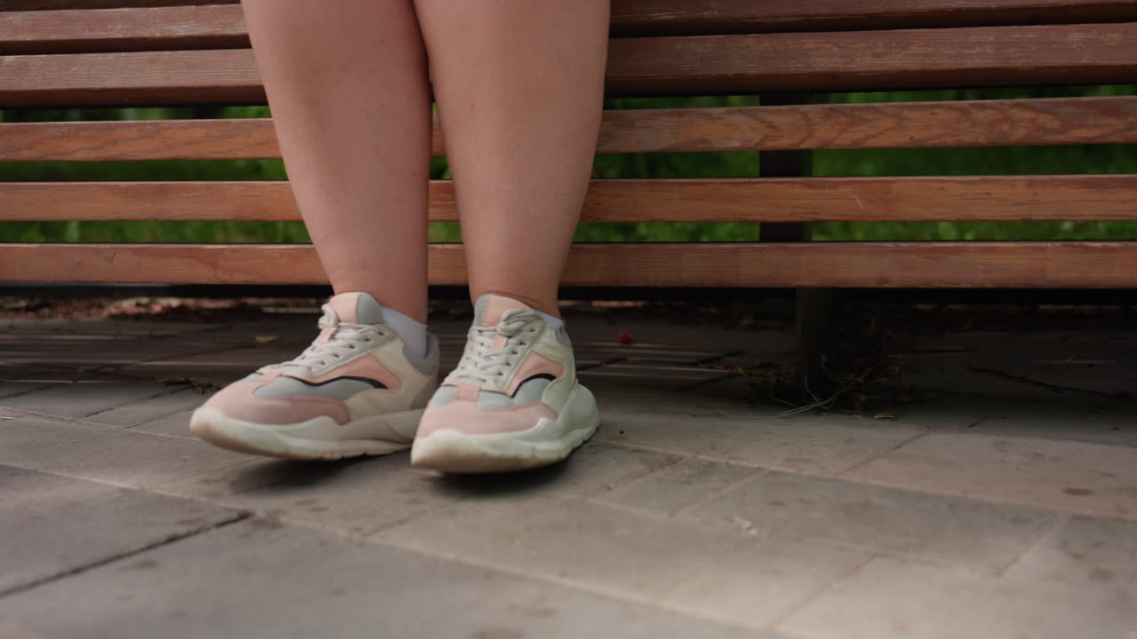 Close up rear view of woman legs in white sneakers walking slowly toward wooden bench in bright park, sun casting soft shadows on pavement, evoking calm atmosphere of leisure and quiet outdoor stroll