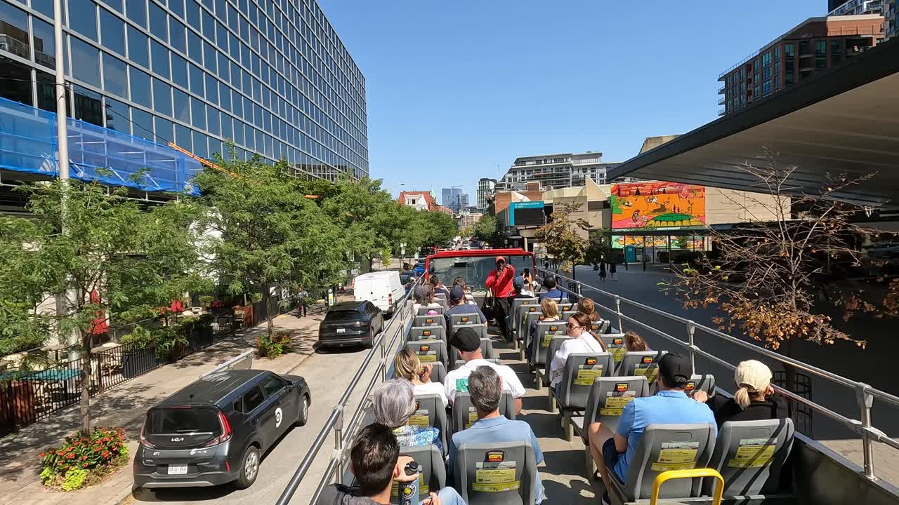 Tourists enjoy a sunny ride on an open-top bus through downtown Toronto, passing modern buildings, colorful murals, and lush greenery, showcasing the city's vibrant atmosphere, Canada