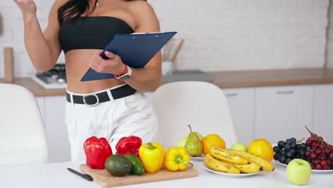 woman with folder in the kitchen. fresh fruit and vegetables on the table. beautiful woman in short top looks at vegetables and writes something. healthy diet.