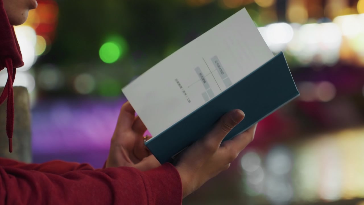 Una persona si ferma a leggere sotto l'illuminazione cittadina. Le luci della città brillano mentre un lettore si concede una pausa per godersi un libro di notte, passeggiando per le strade illuminate