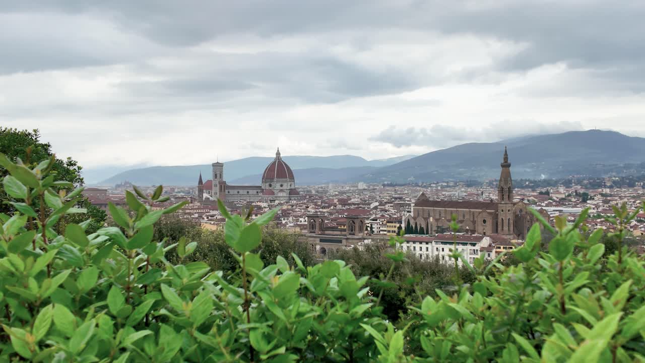 Wide angle establishing shot of Florence, Italy, featuring the iconic Duomo and city skyline beyond lush green foreground leaves under dramatic cloudy skies, soft natural light