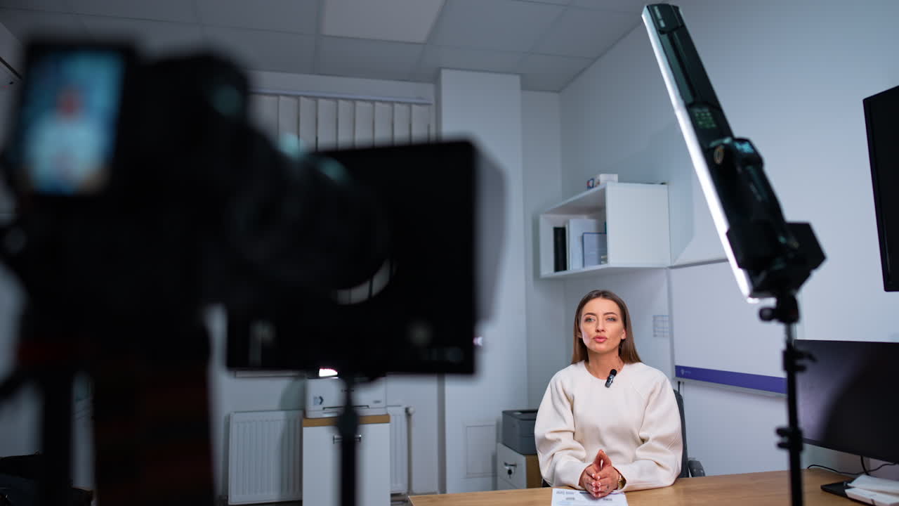 Female blogger influencer sitting at desk talks to camera. Blurred view of a camera and a lamp at foreground.